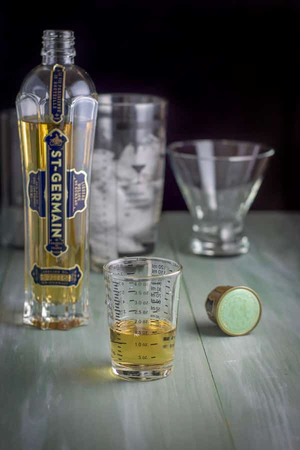 Elderberry liqueur measured with the bottle, shaker and glasses in the background