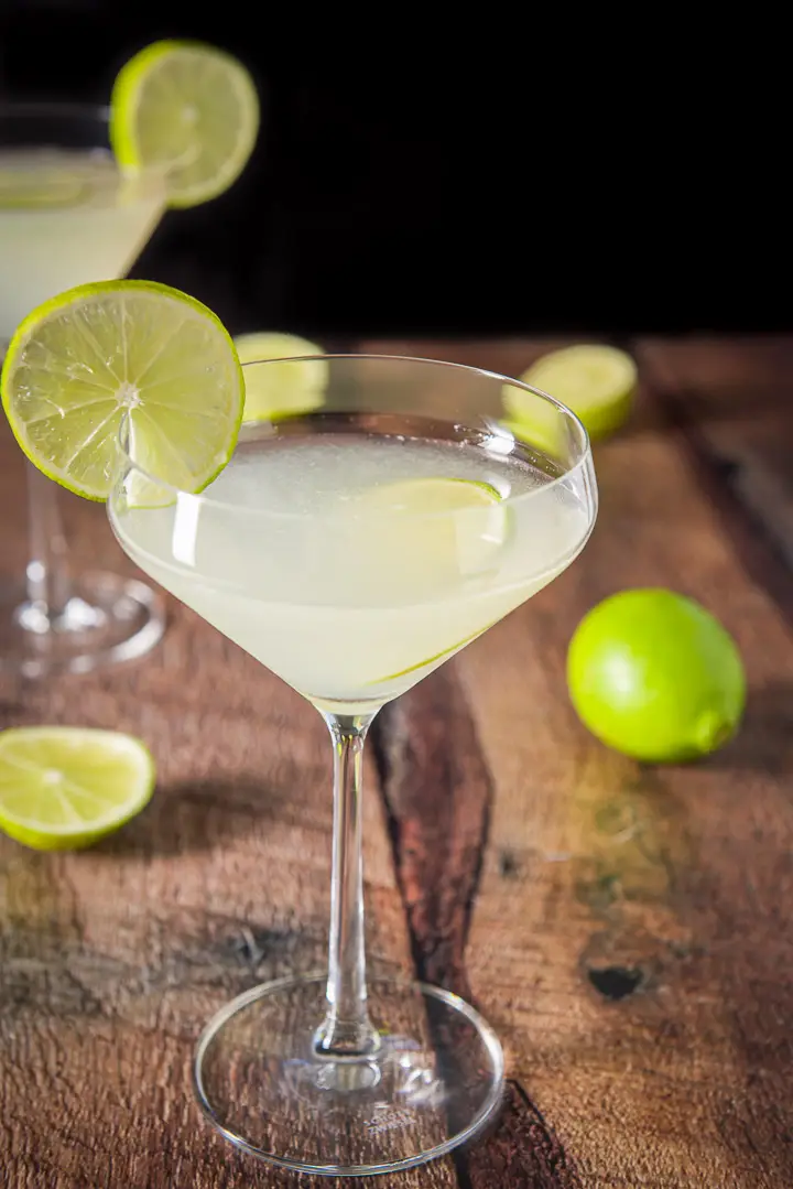A curved martini glass with lime liquid garnished with lime wheels. There are also limes on the table and another glass in the background
