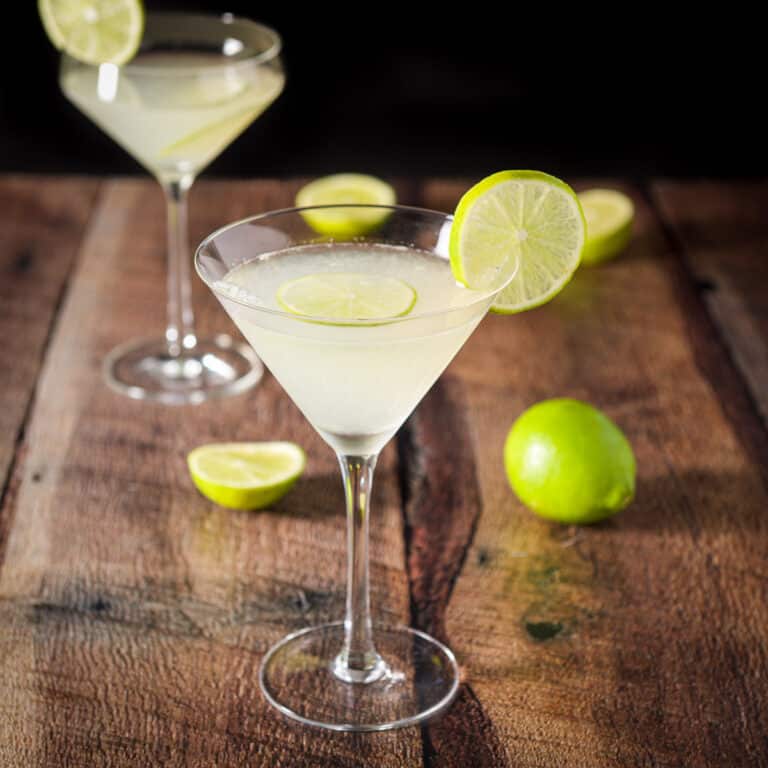 A traditional martini glass filled with a lime drink in front of a curved glass. There are limes on the side of the glasses and in them as well as on the table - square