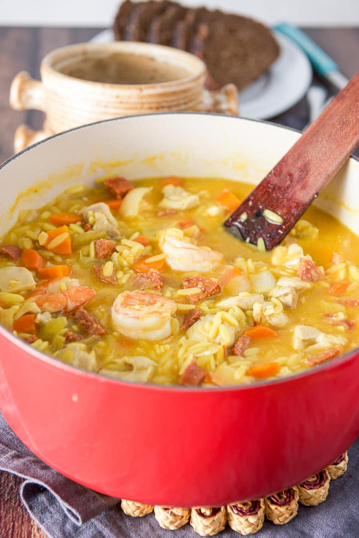 A red Dutch oven filled with the soup filled with shrimp, chicken and chorizo. There is crocks and bread in the background