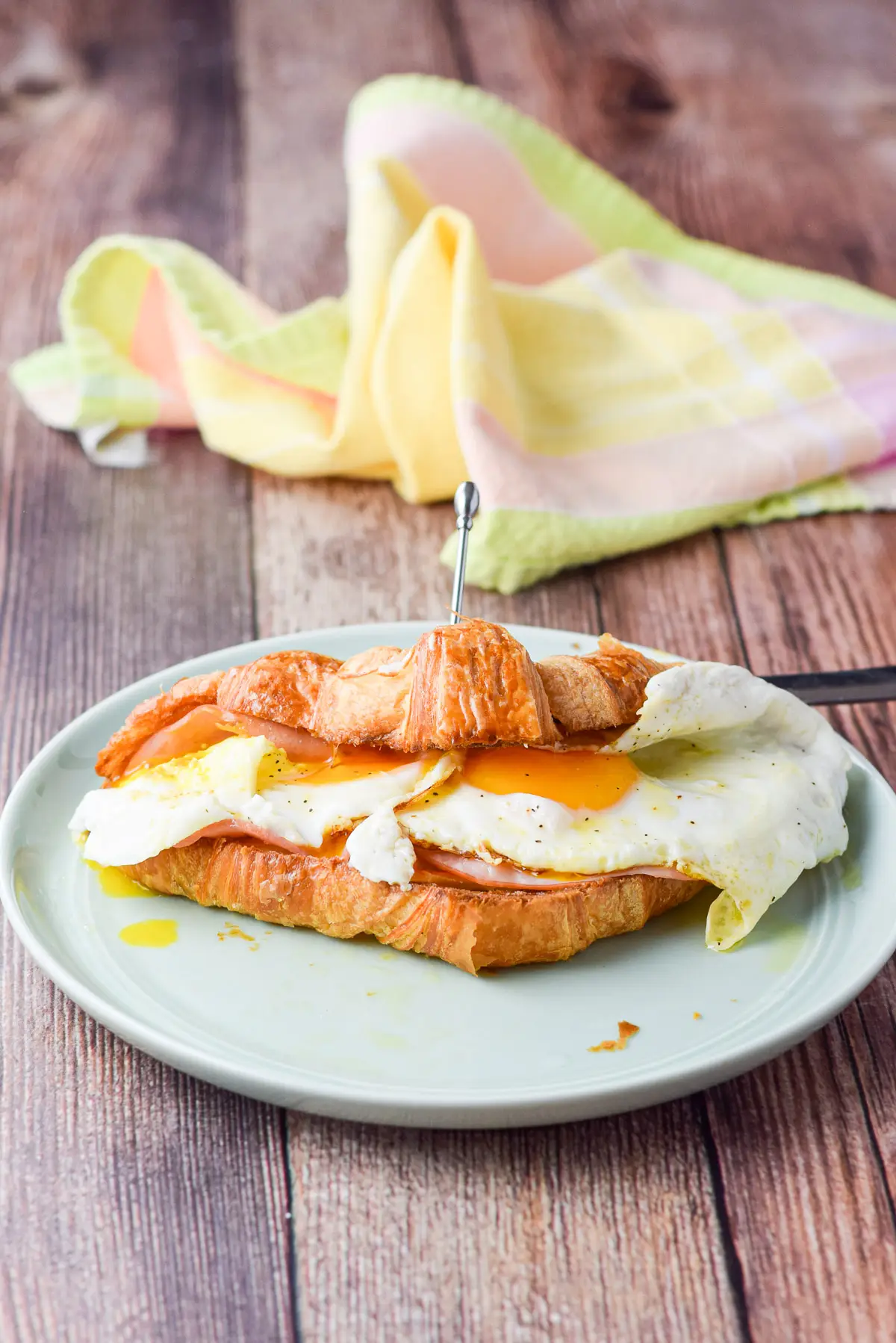 Eggs on a croissant with ham and cheese. There is a pick holding the croissant closed