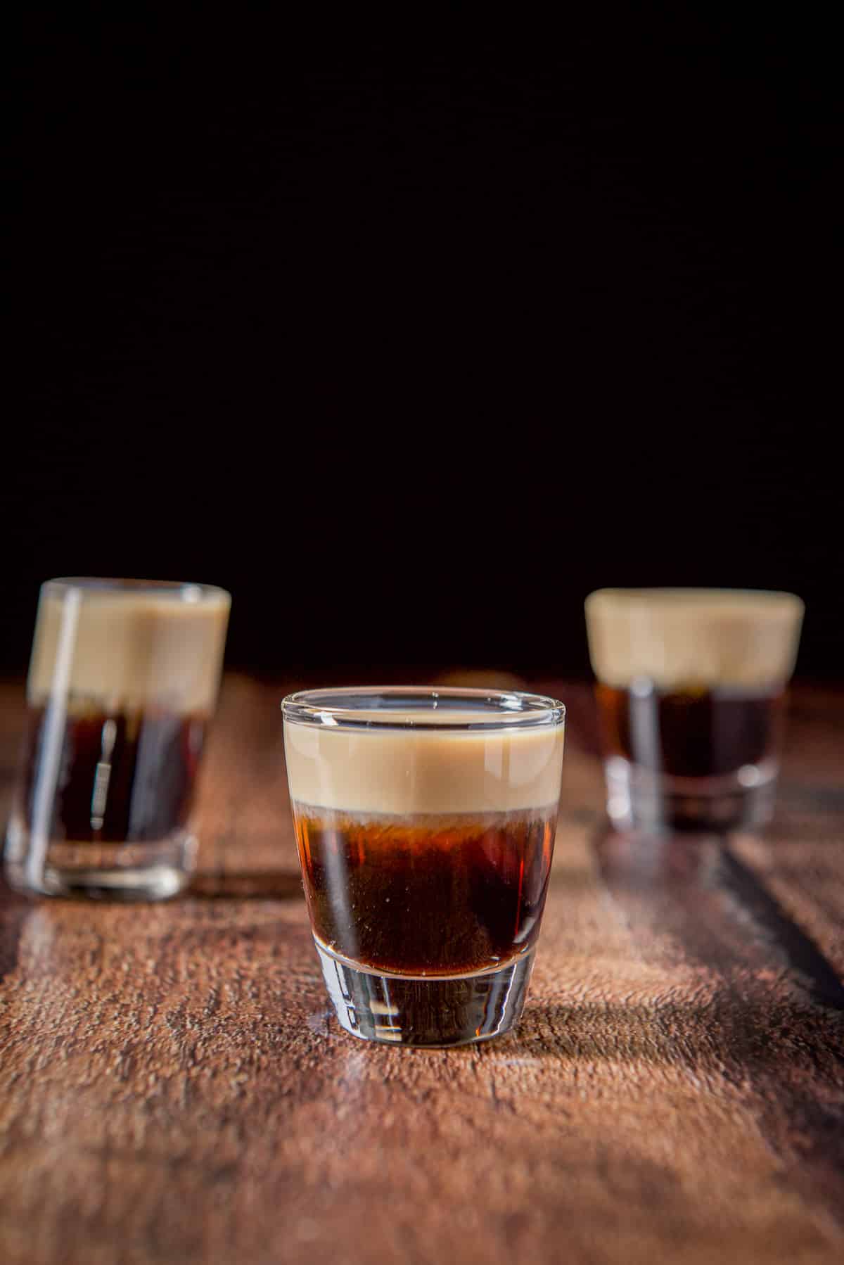 The short glass filled with the layered shot on the wooden table. There are two other glasses behind it