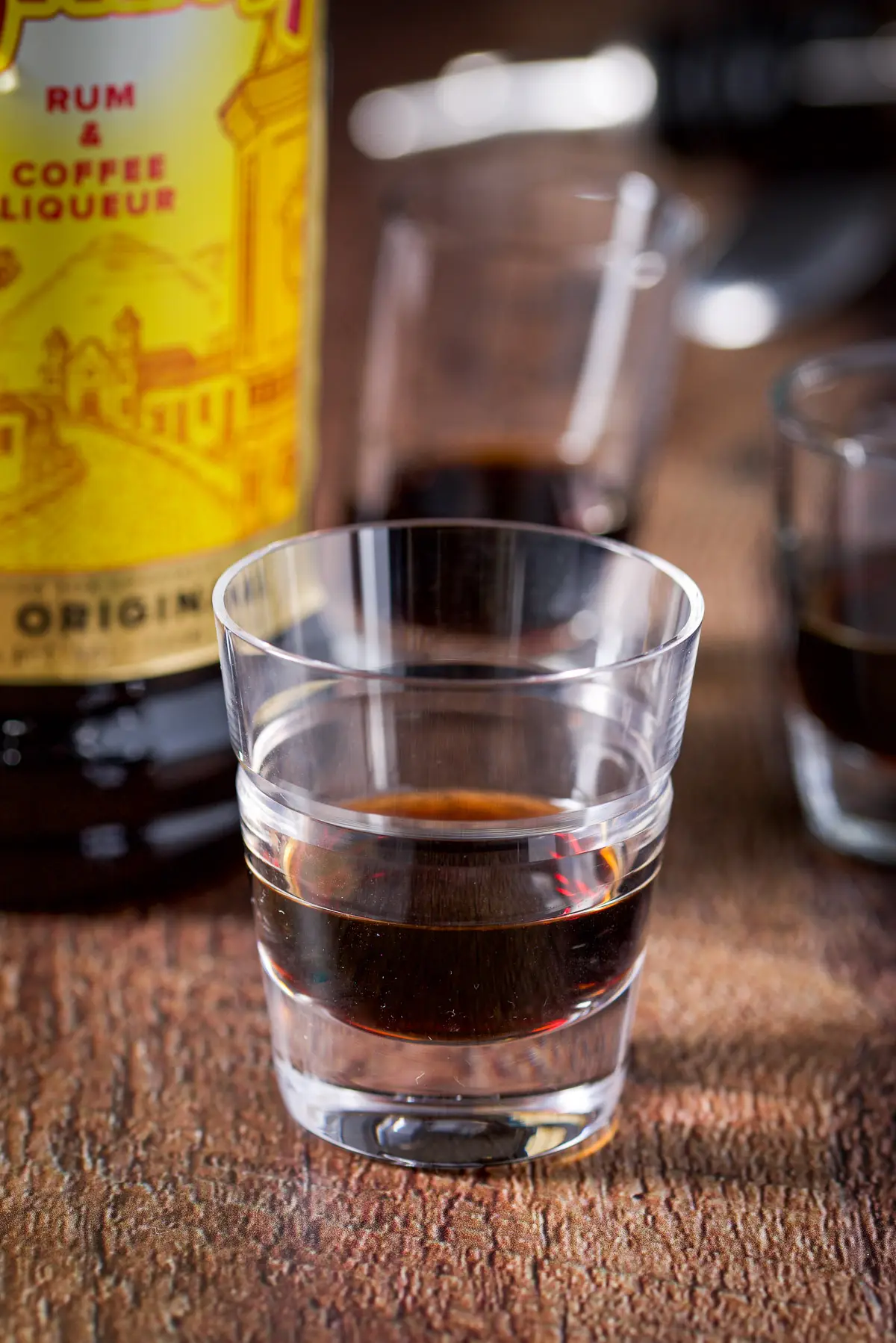 A beveled glass in front with Kahlua in it with the other glasses and bottles in the background