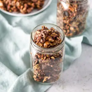 Chocolate granola in a glass jar with another jar and plate in the background - square