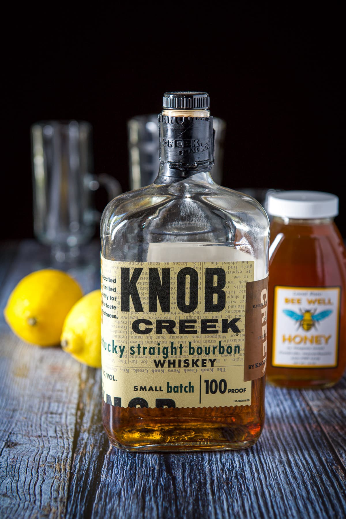 Bourbon, honey, lemon on the grey table with a shaker and glassware in the background