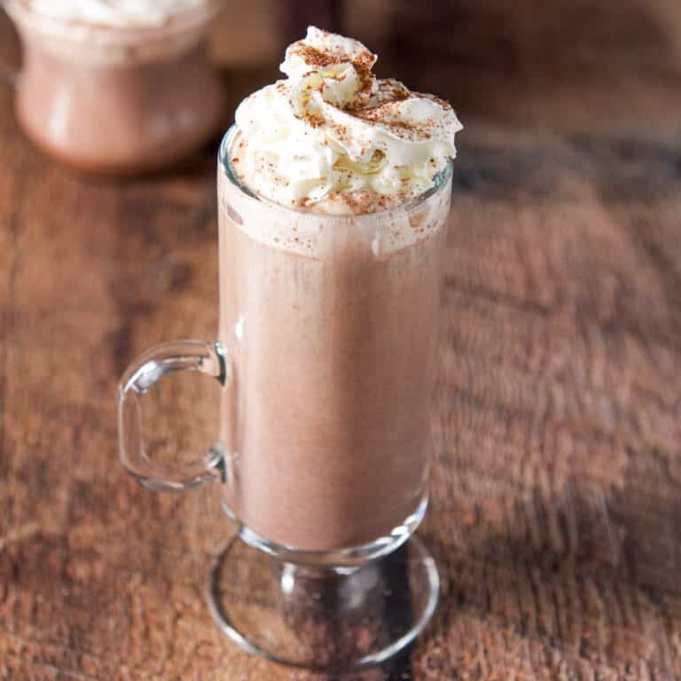 A clear mug with a hot chocolate drink with whipped cream on top - square