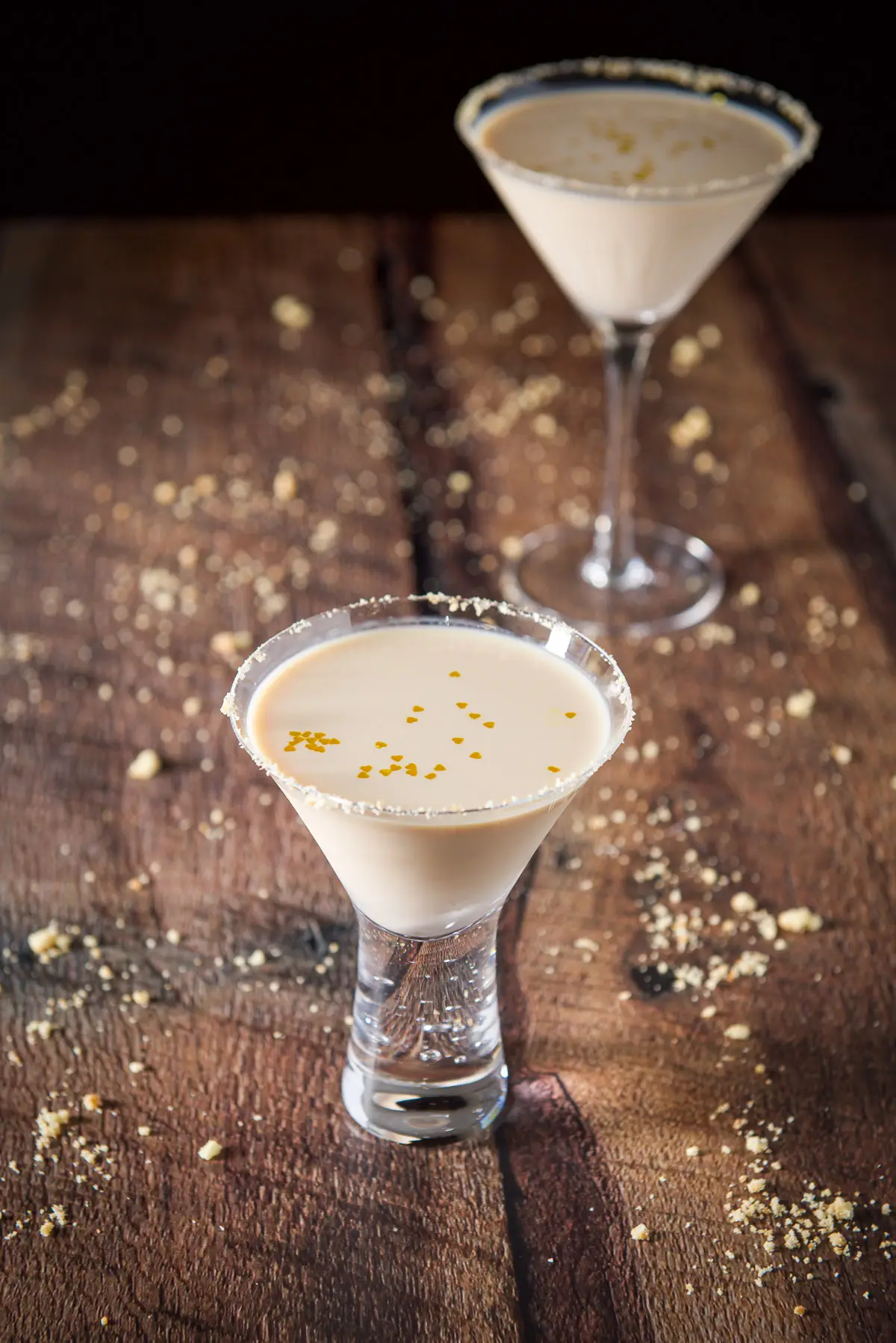 A short bubble glass with the cream drink with gold hearts floating in it. There is another glass and cookie crumbs on the table