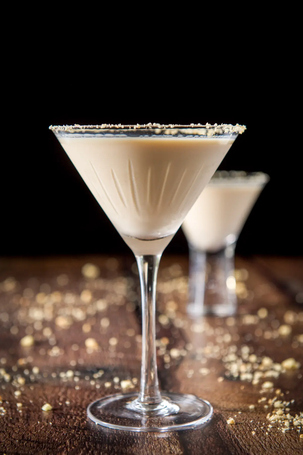 Vertical view of the classic martini glass in front of the smaller glass with the cream drink. There are cookie crumbs on the table