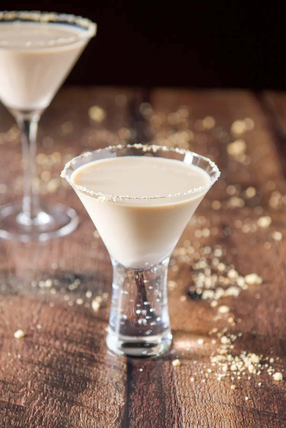 A small bubble glass garnished with cookie crumbs and the cream drink in it. There is a classic martini glass and cookie crumbs on the table behind it