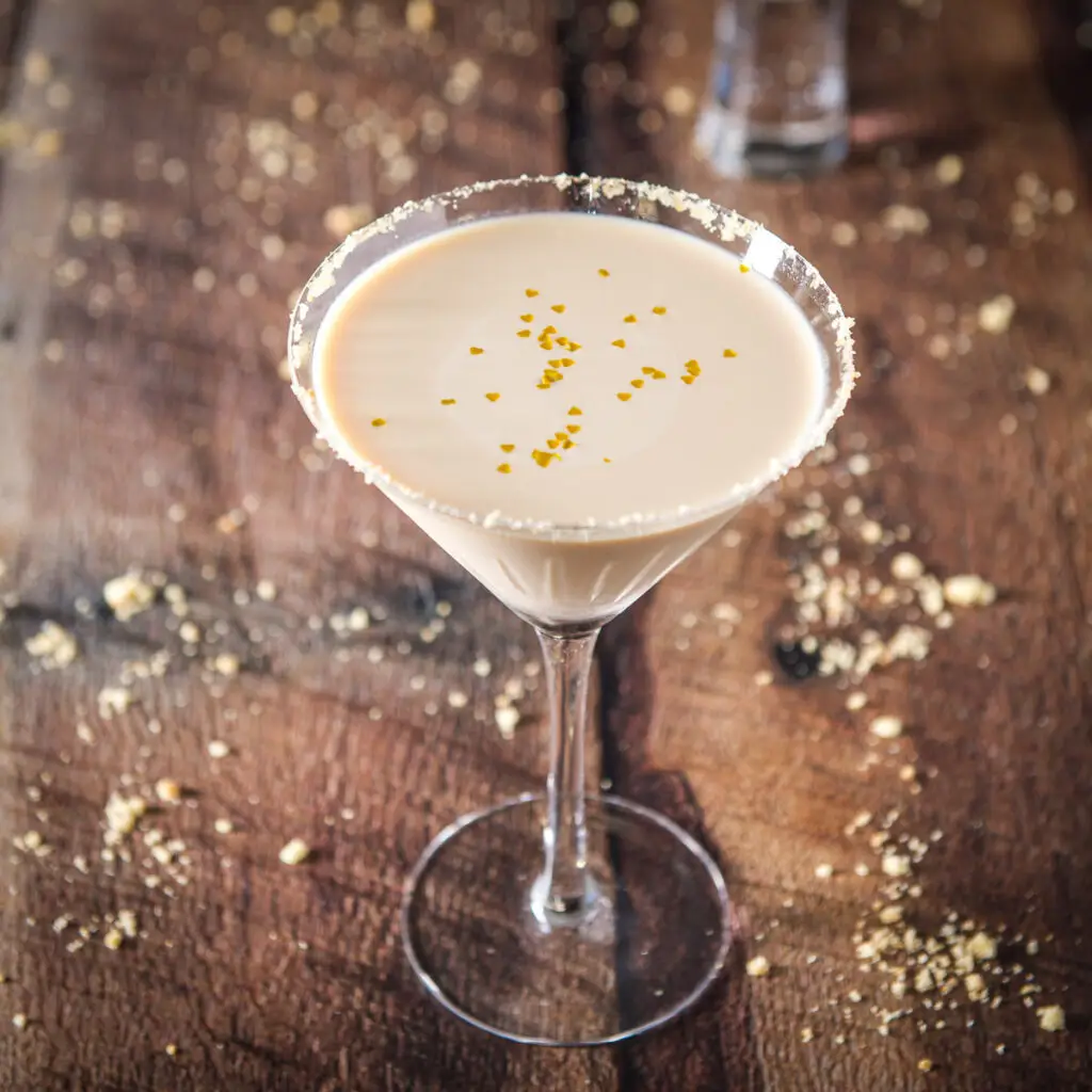 A classic martini glass garnished around the rim with sugar cookie crumbs. Inside the glass is a cream drink with little gold hearts floating - square