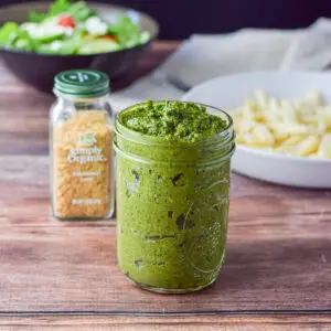 A jar of pesto sauce with yeast, salad and a bowl of pasta in the background
