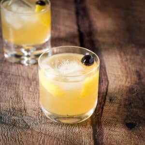 Two classes with golden cocktails with lemon twists and maraschino cherries - square