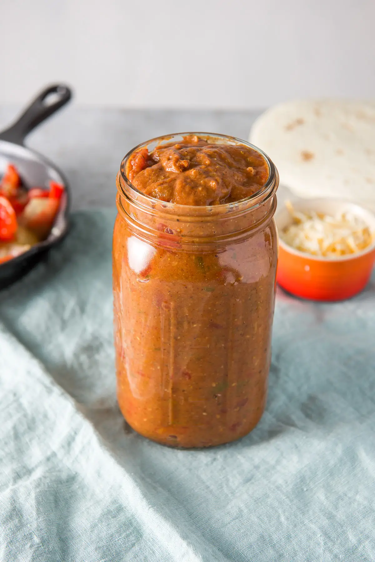 A jar of the red enchilada sauce and vegetables in a pan and tortillas and cheese in the background