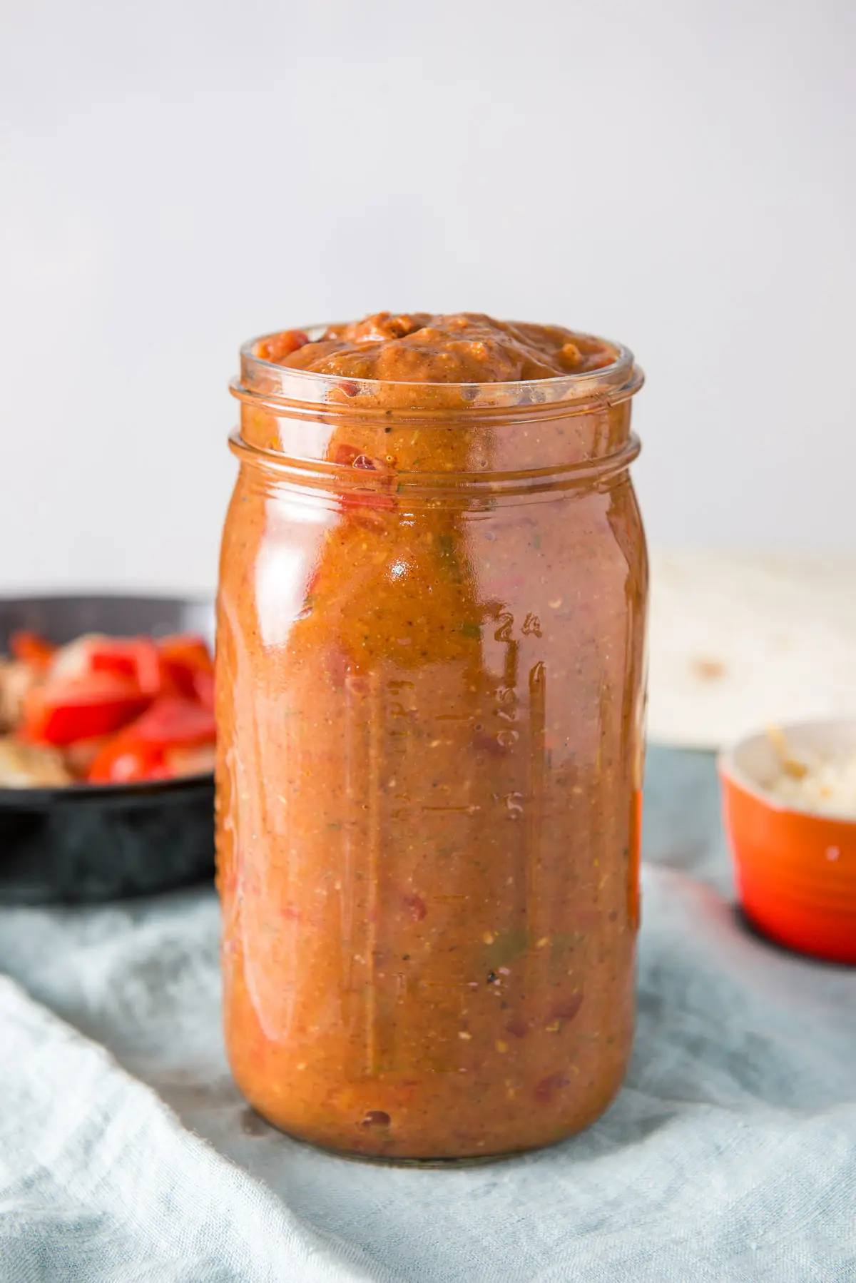 Vertical view of a large jar filled with the red sauce. There is a pan of vegetables, tortillas and cheese in the background