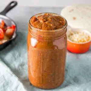 Jar filled with the enchilada sauce with tortillas, cheese and veggies in the background