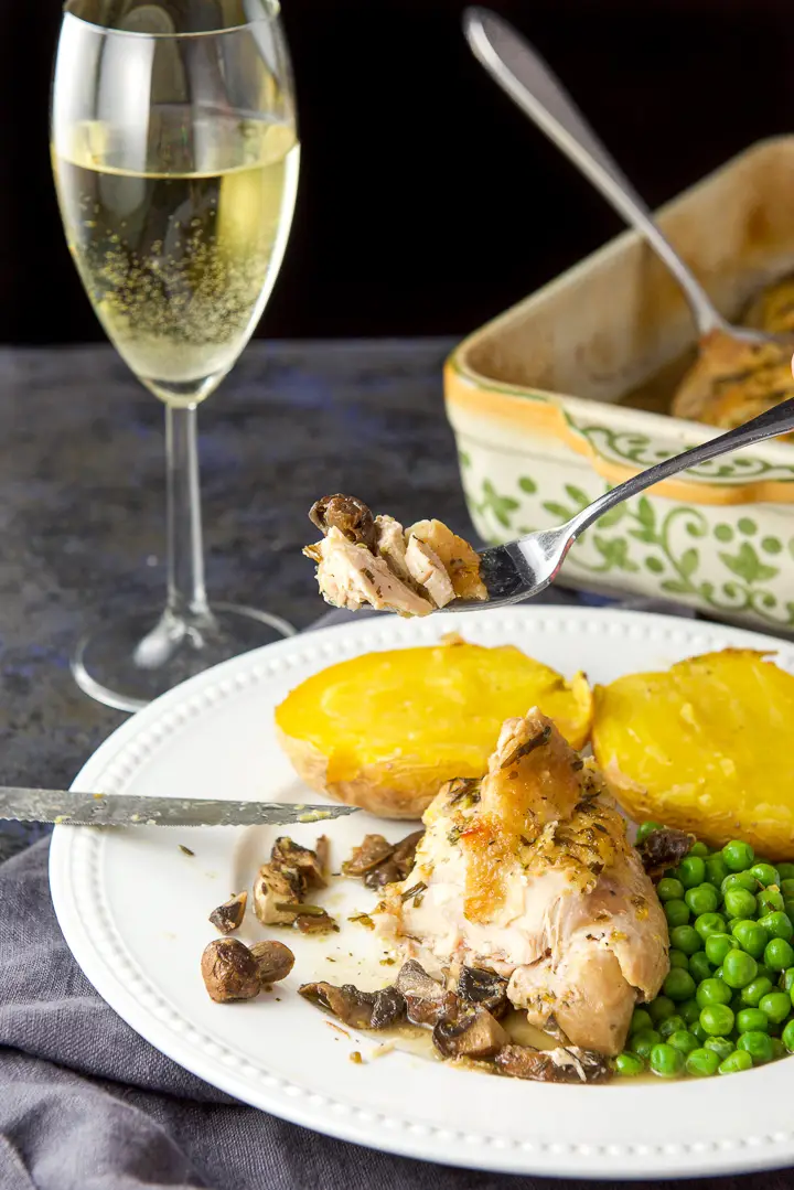 A fork with chicken on it held over the plate with wine and the baking dish held in the background