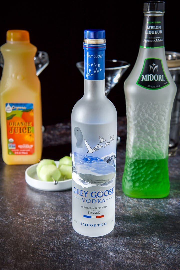 Vodka, Midori, orange juice and melon on a pick with glasses and shaker in the background