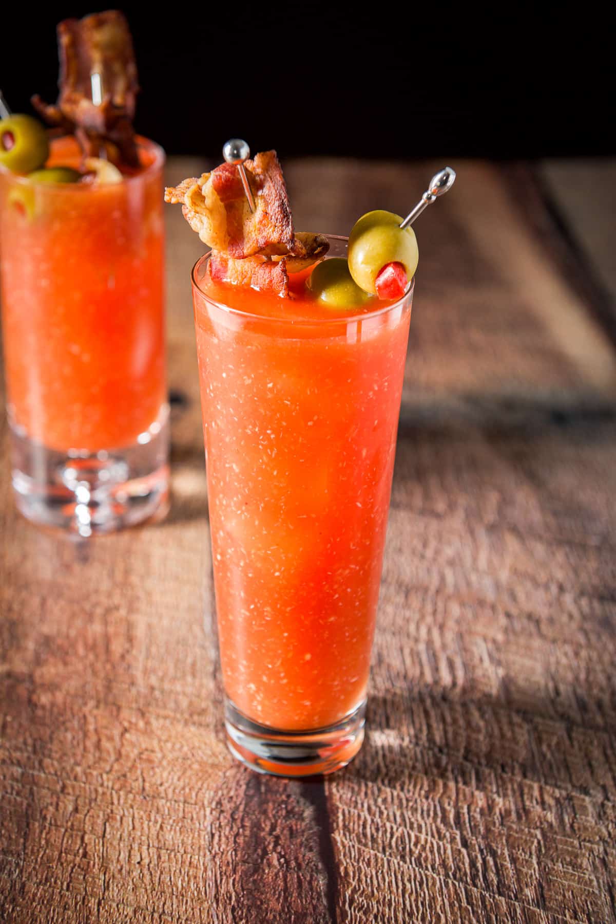 Olives and slices of bacon on picks in two glasses filled with the cocktail