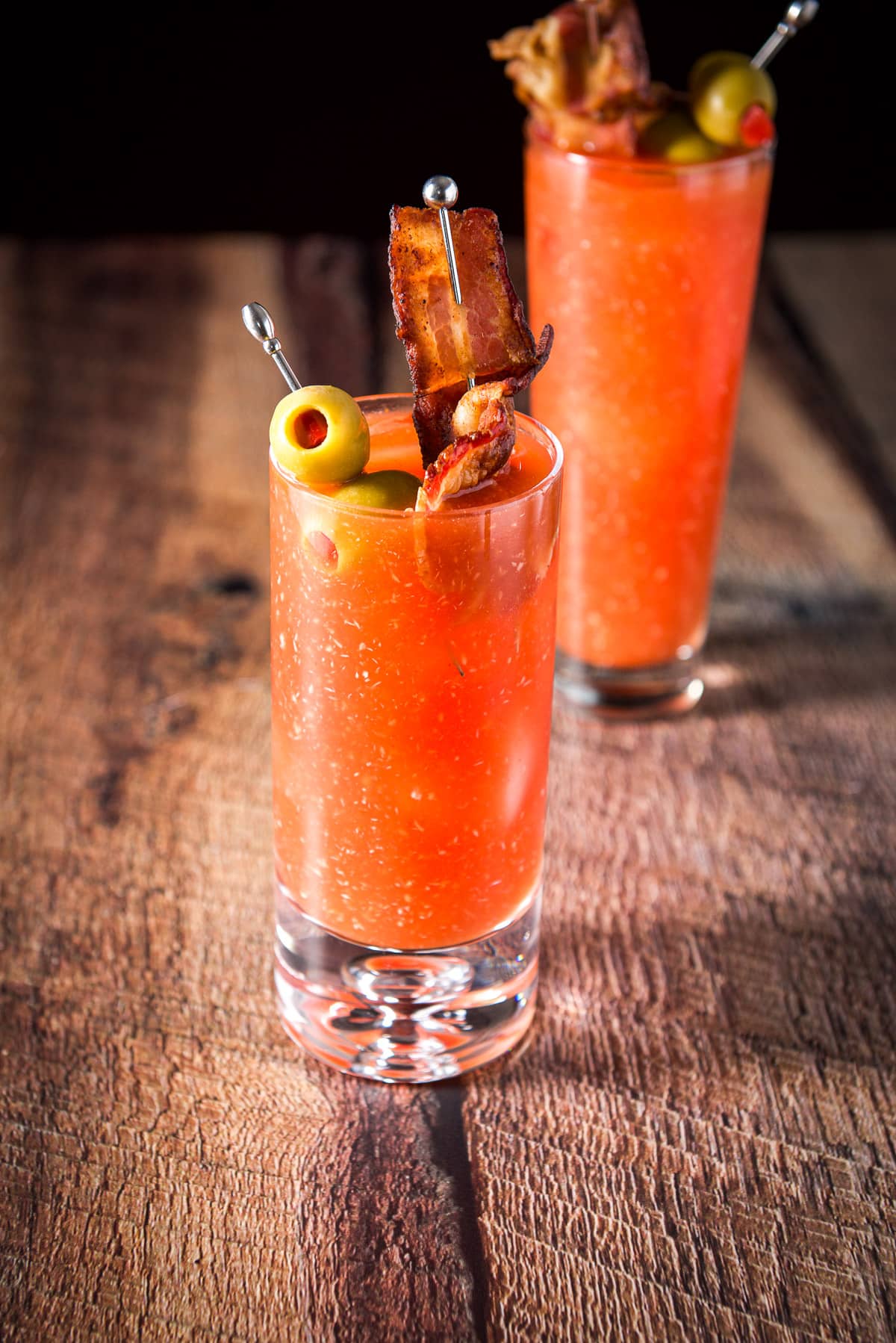 A rocks glass filled with the tomato cocktail with two picks of garnish in front of a flared glass