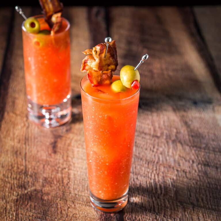 A tall flared glass filled with the red cocktail with horseradish floating. There are olives and a piece of bacon as garnish - square