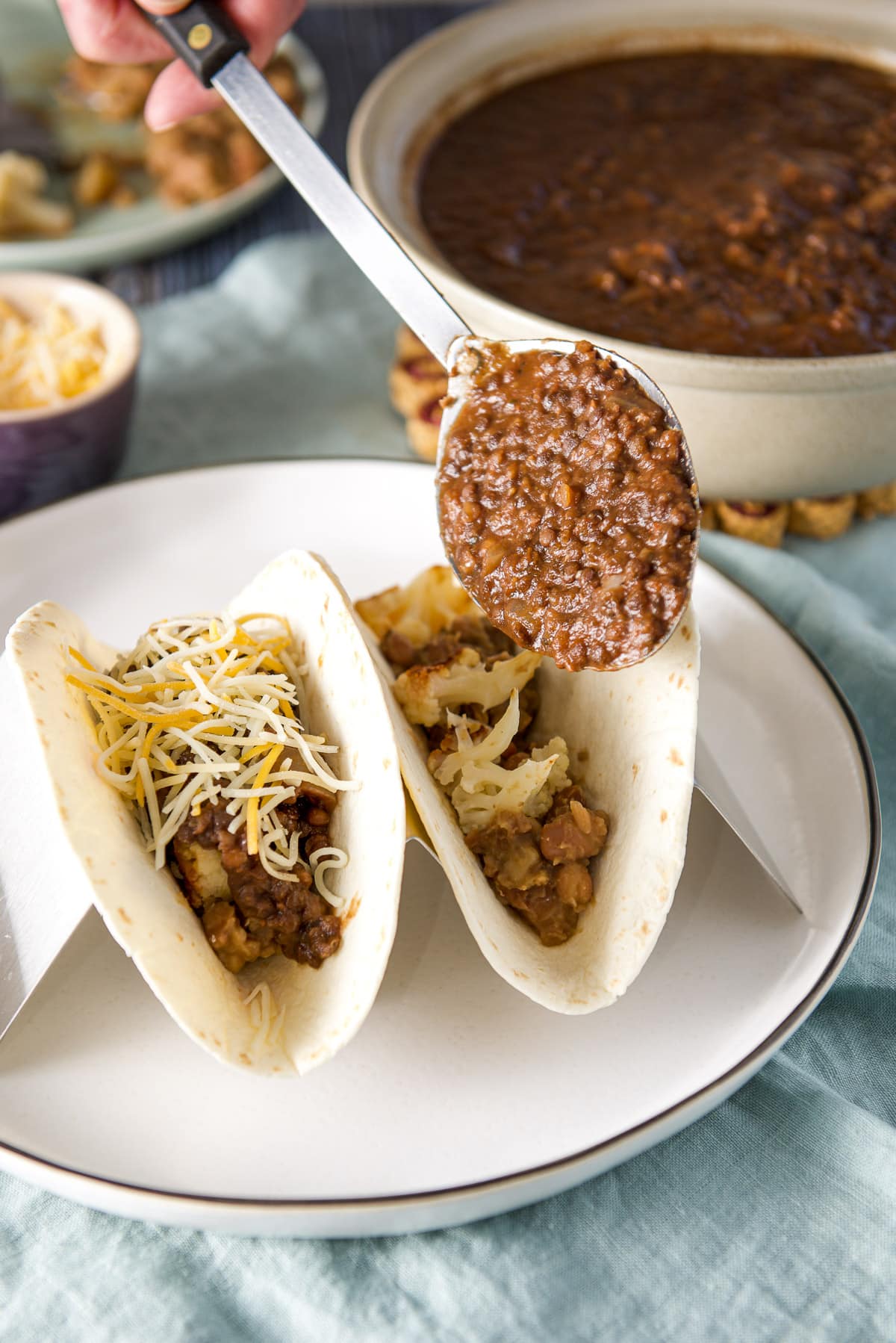 A large spoon of lentils held over the two flour tortillas with a big bowl in the background
