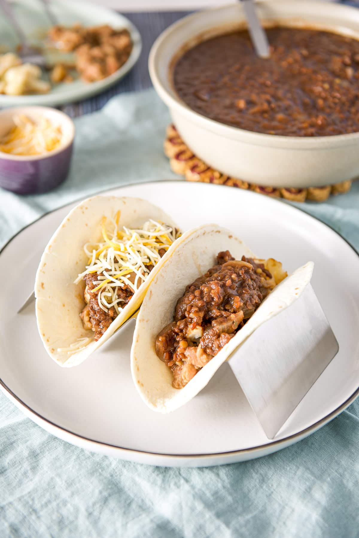 A white plate with a taco holder with flour tortillas and the taco filling in it along with a bowl of lentils in the back