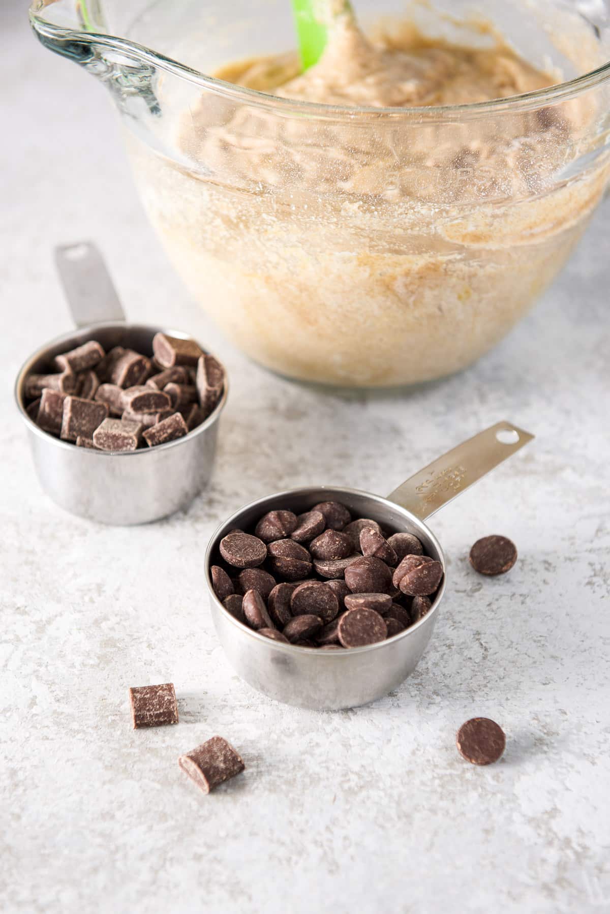 Two measuring cups with big chocolate ships and square chips with a bowl of batter in the background