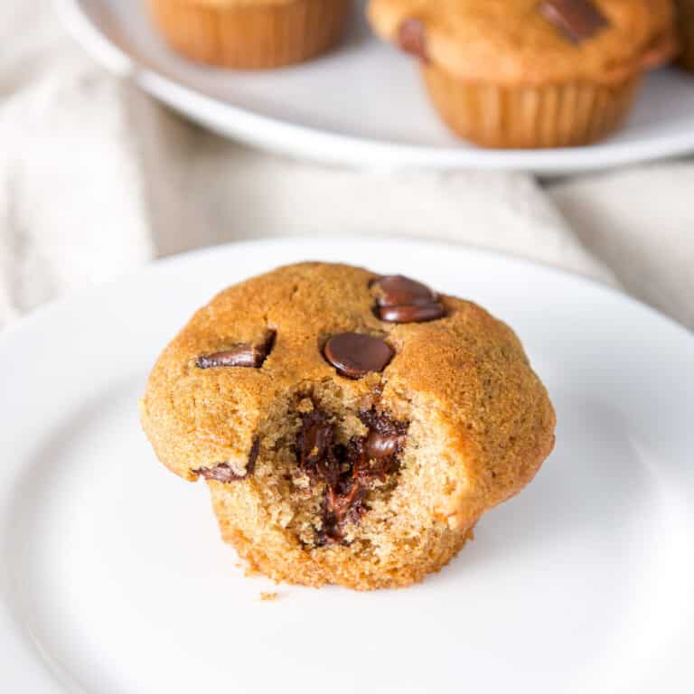 Close up of a bite taken out of a chocolate chip muffin on a white plate with more in the background - square