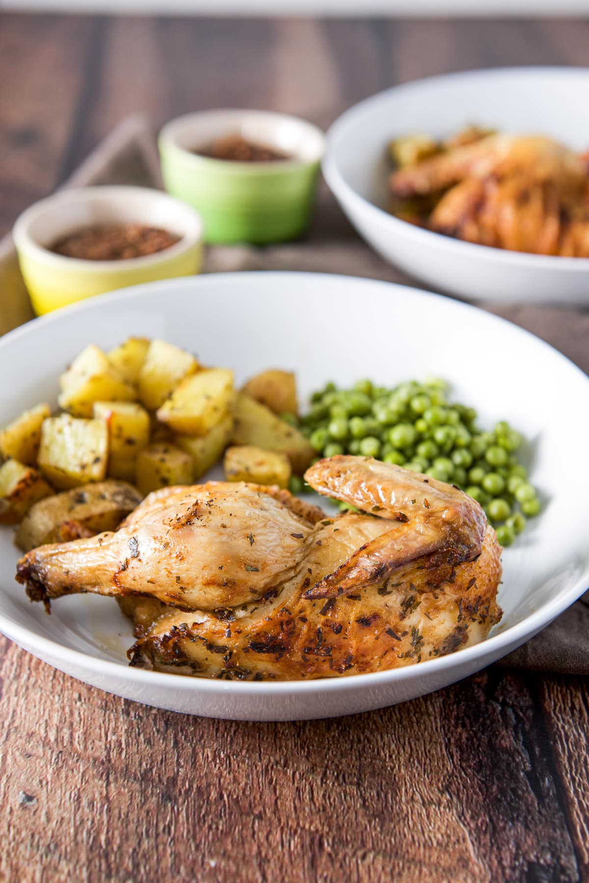 Vertical view of half a hen on a white plate with peas and potatoes. There is another plate in the background with ramkens of lentils