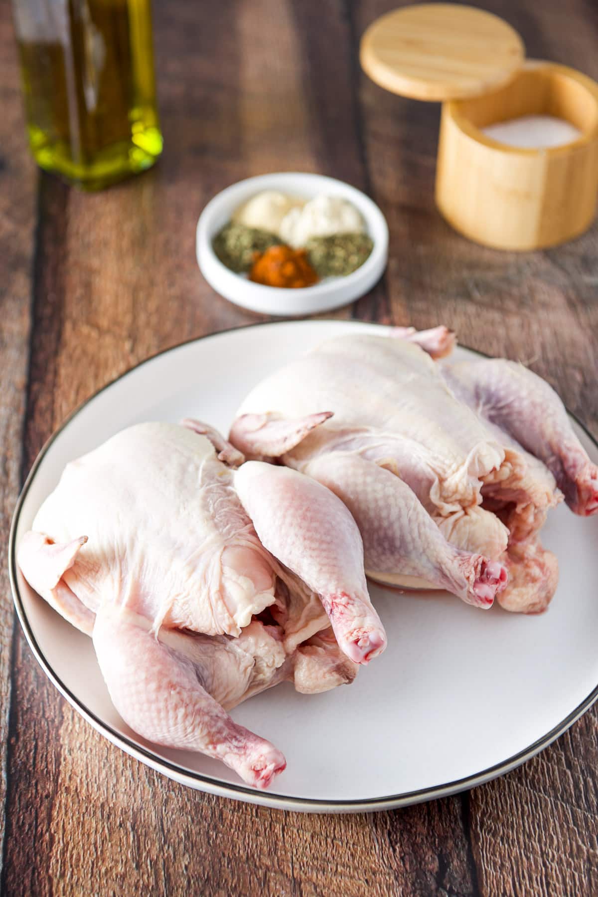 A white plate with two hens on it, with a white plate of herbs and spices, salt and olive oil in a bottle