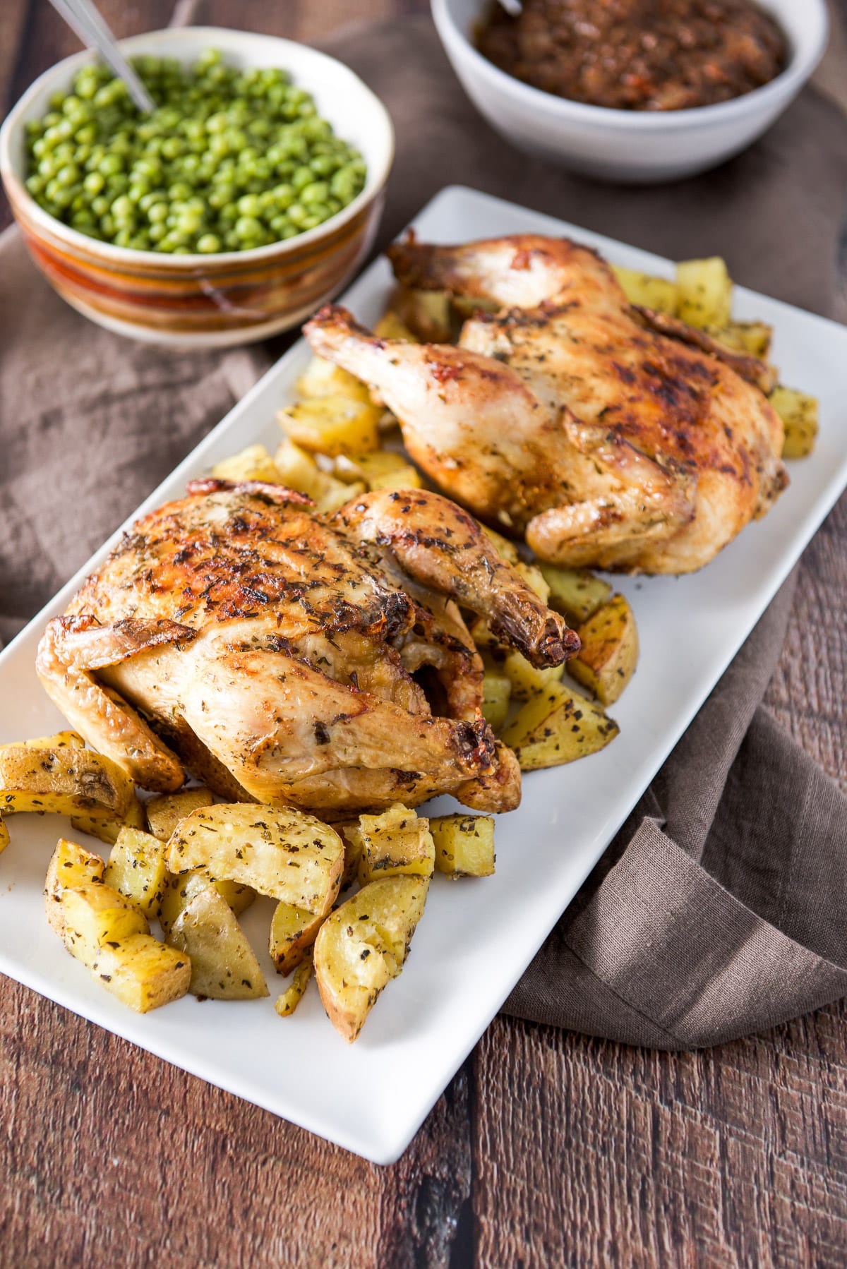 A white platter with roasted potatoes and two cooked small hens with peas and lentils in the background