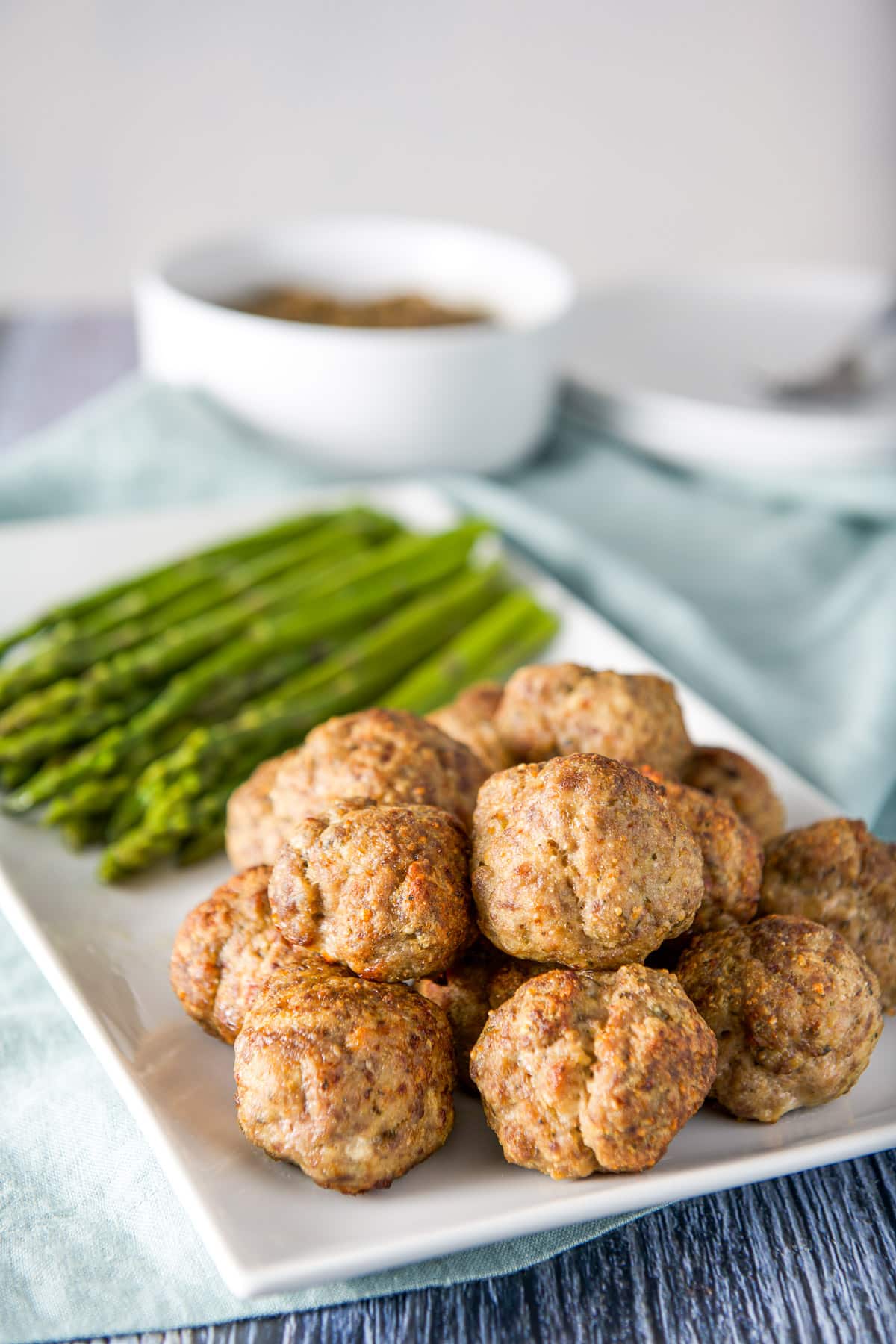 A white platter with meatballs stacked on each other, asparagus and a bowl of lentils in the back