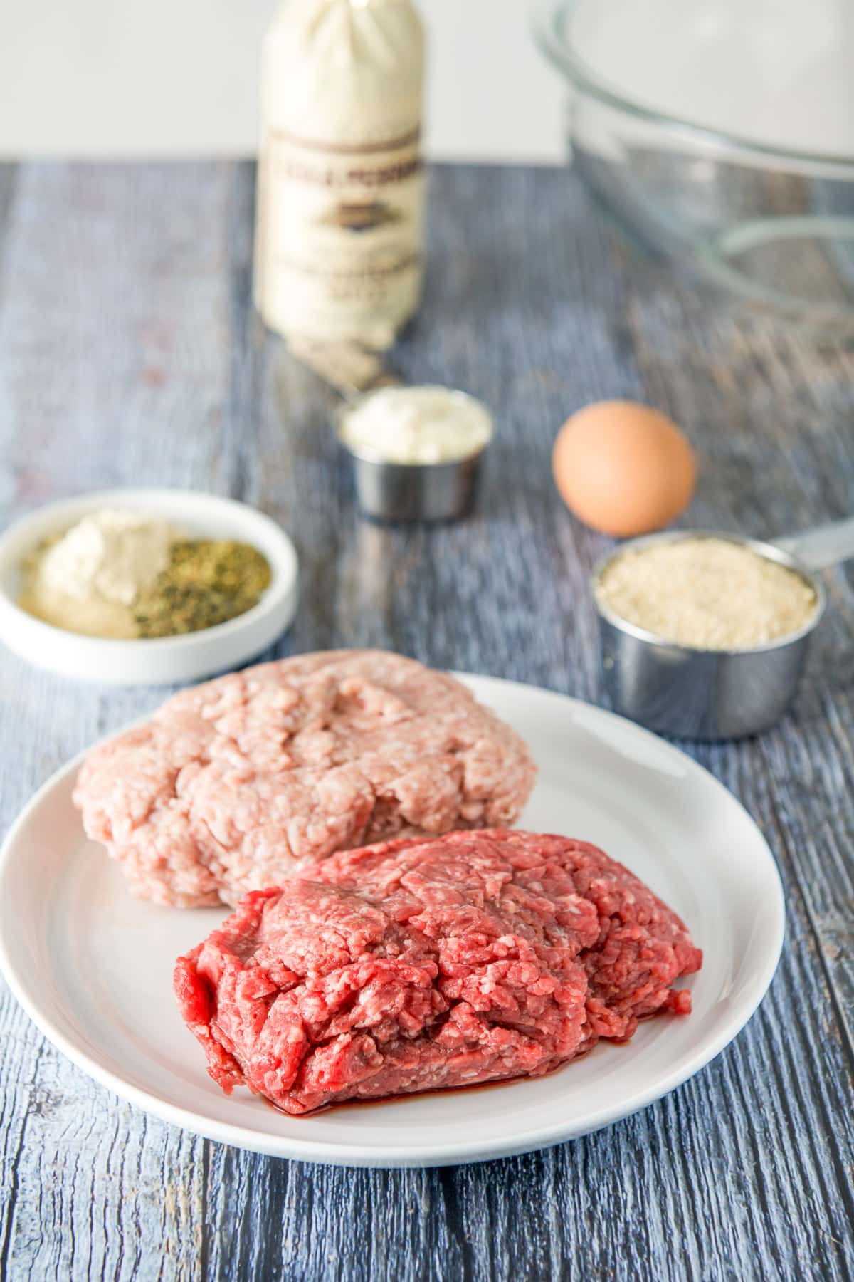 A white plate with ground beef and pork with spices, herbs, parm cheese and an egg in the back