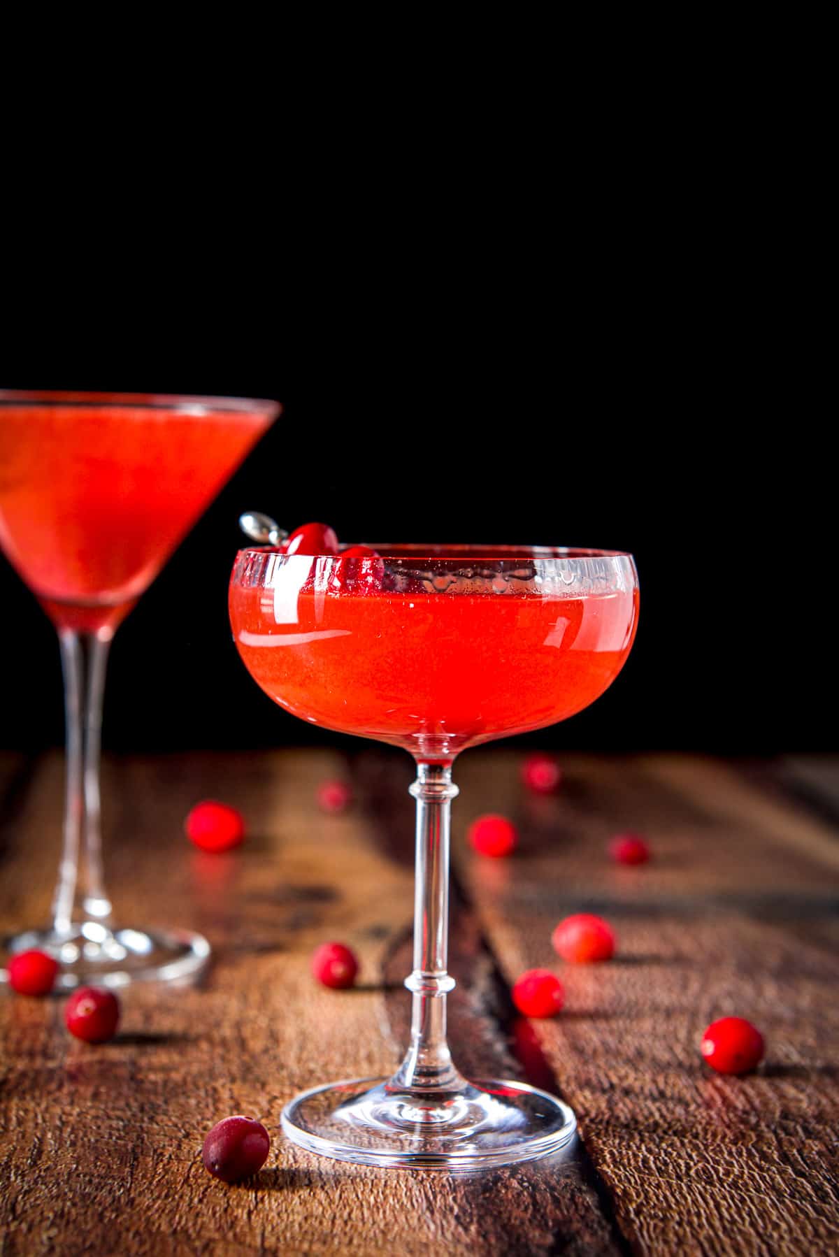 Vertical view of the beveled coup with the cranberry drink with cranberries strewn on the table and another martini glass in the background