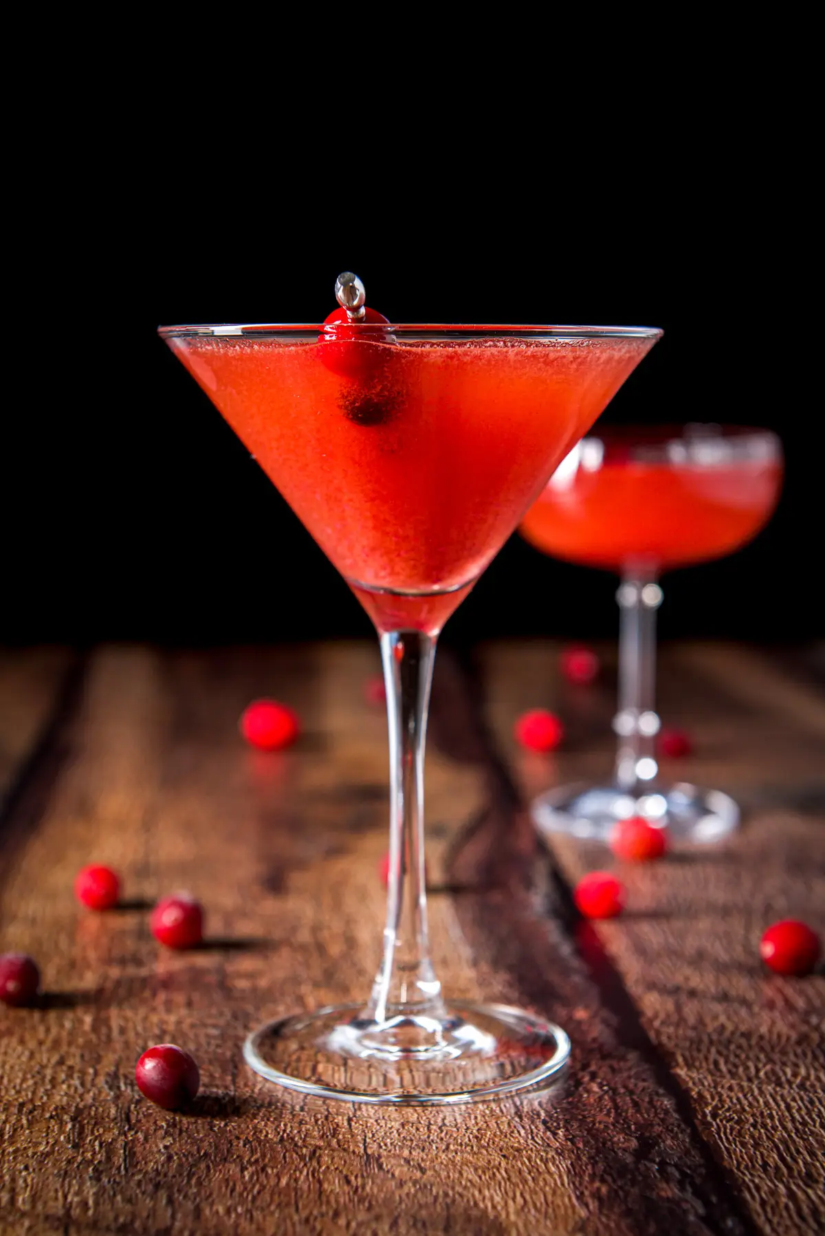 Classic martini glass with the red cocktail in it along with cranberries on a pick