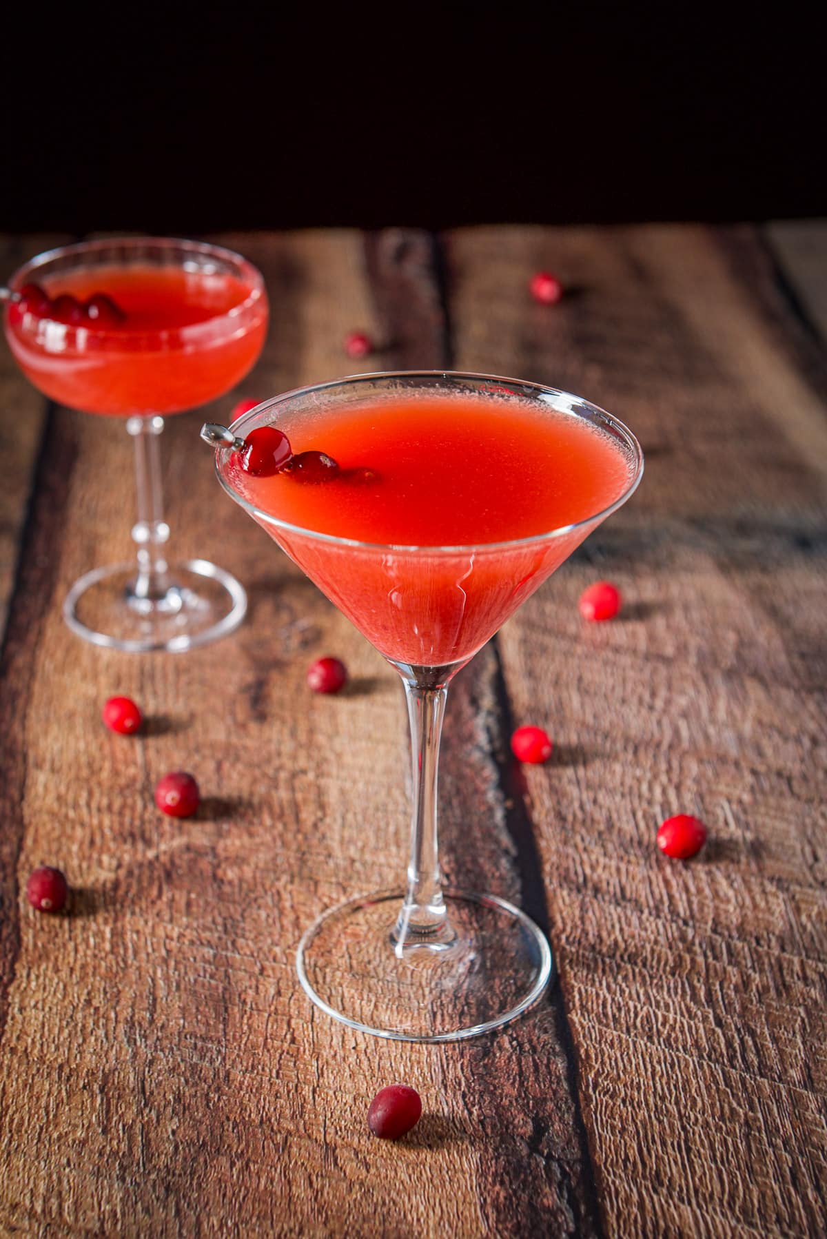 Classic martini glass in the front of the coupe glass with the cranberry cocktail and cranberries strewn on the table