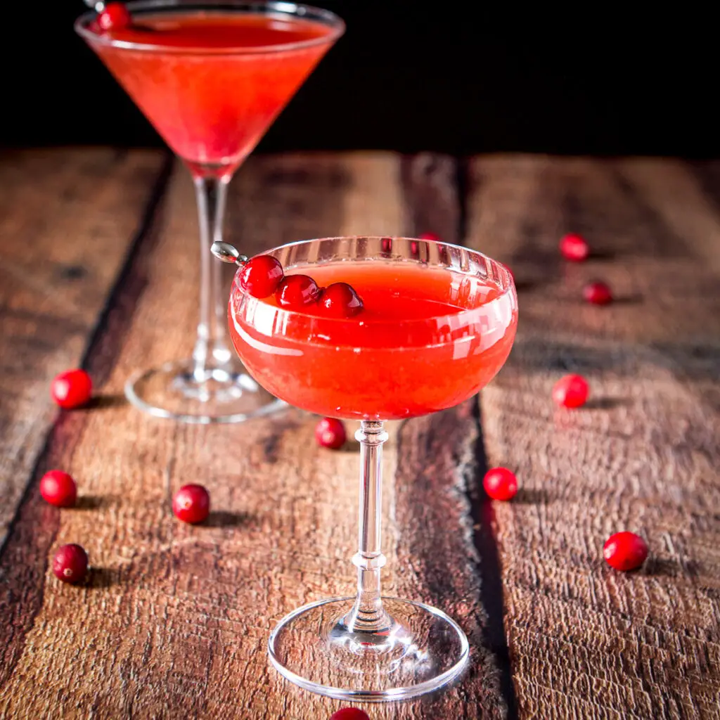 A beveled coupe glass filled with a cranberry cocktail. there are cranberries on the table and a classic martini glass in the back - square