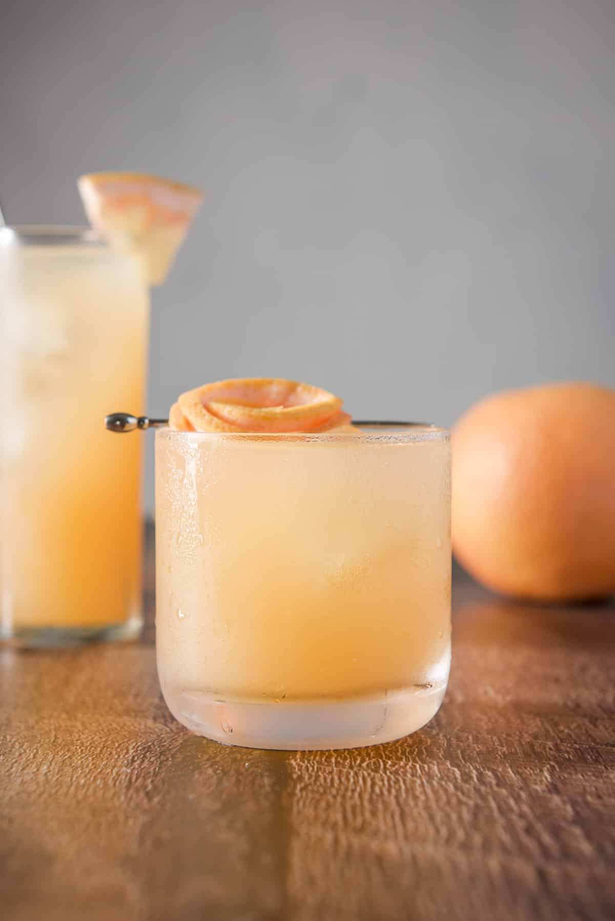 Vertical view of the cocktail in the short glass in front of the collins glass. Both are garnished and there is a grapefruit in the background