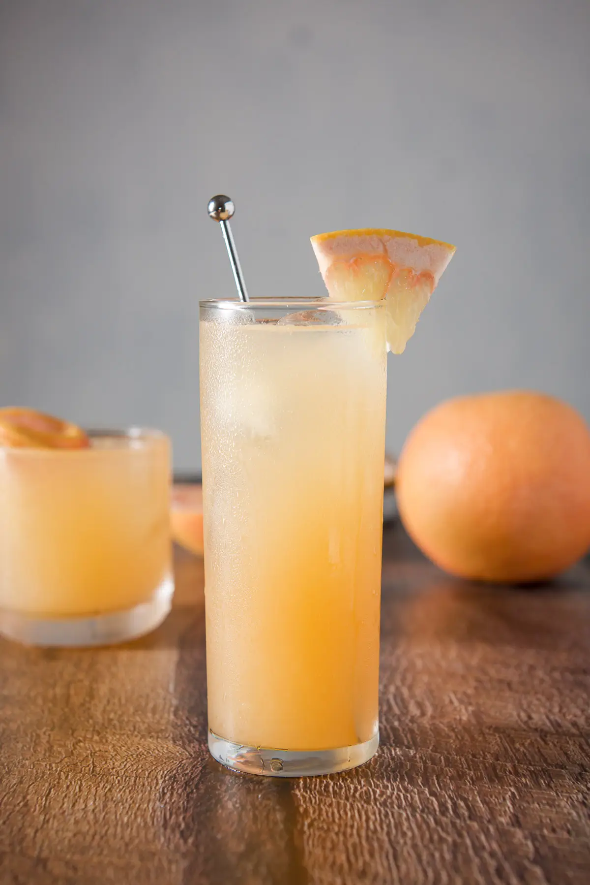 Vertical view of the tall collins glass with the cocktail with a stirrer and wedge of grapefruit. There is a shorter glass in the background and a whole grapefruit