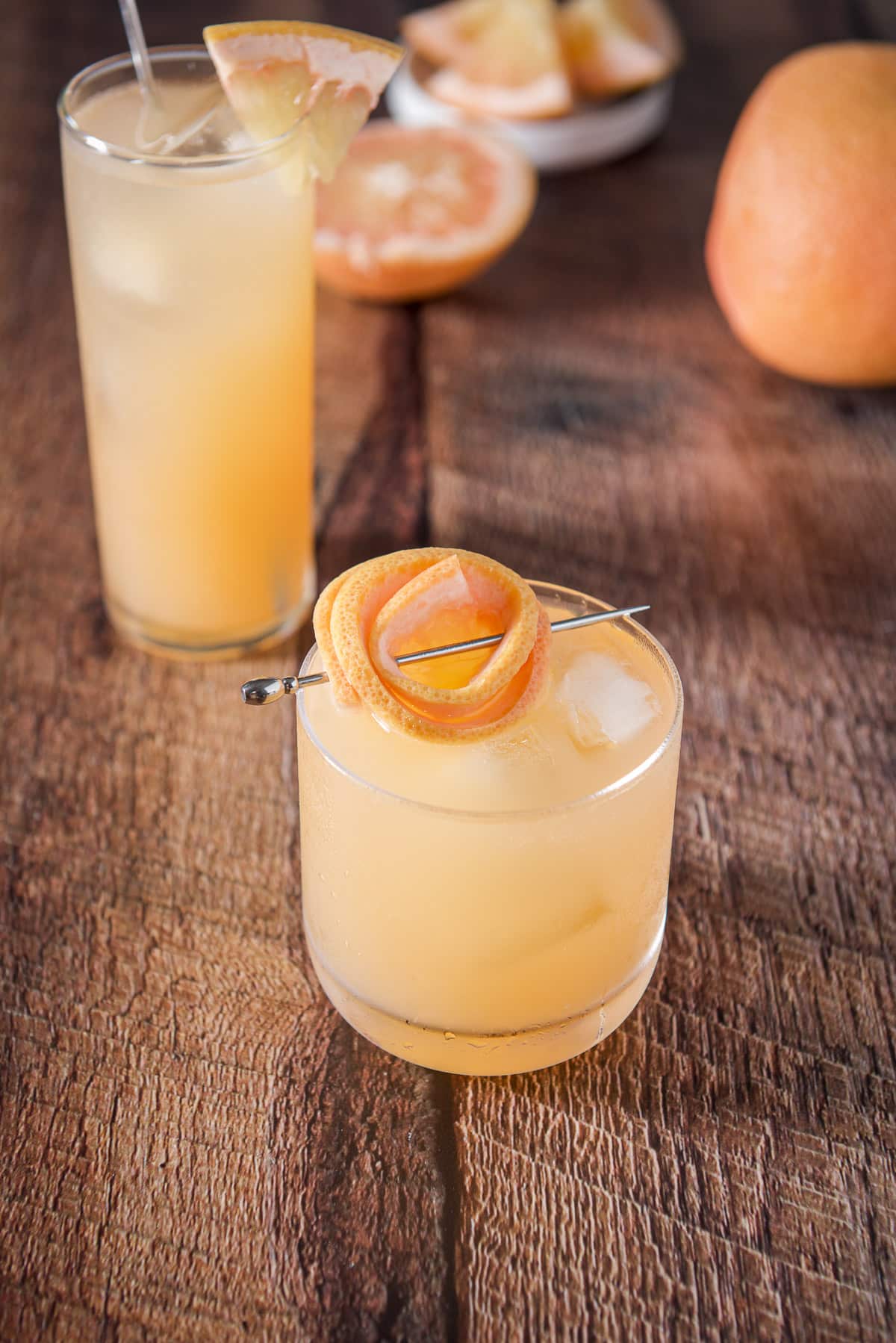 A double old fashioned glass and tall glass filled with the grapefruit cocktail, both have garnishes of grapefruit
