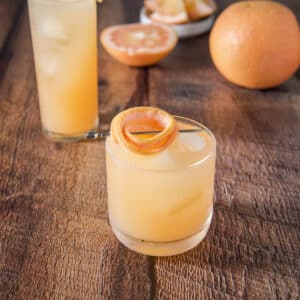 Glass with a grapefruit garnish on the side of the glass with a big grapefruit and slices in the background - square