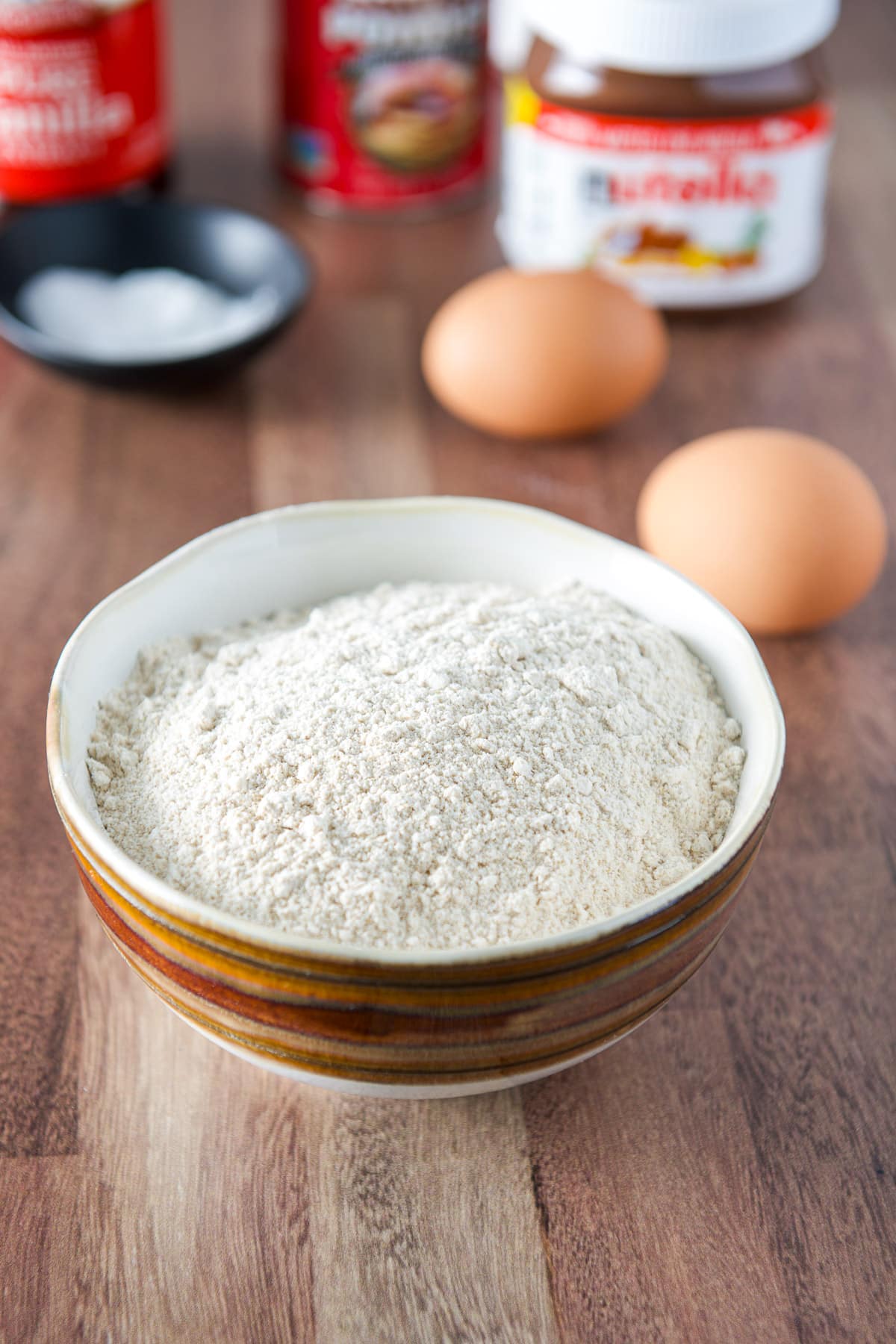 A brown bowl of flour, two eggs, Nutella, baking soda and powder