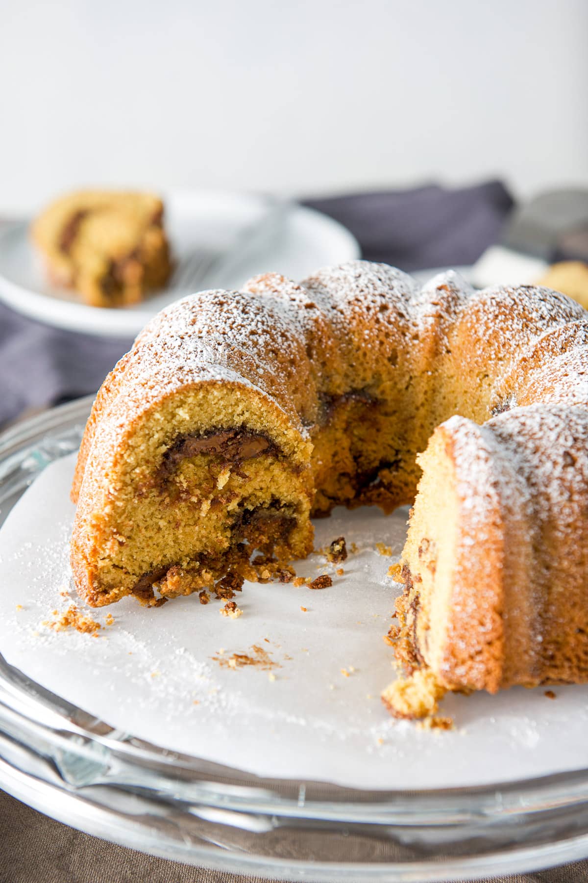 A coffee cake with pieces taken out of it on a cake stand with a plate with a piece in the back