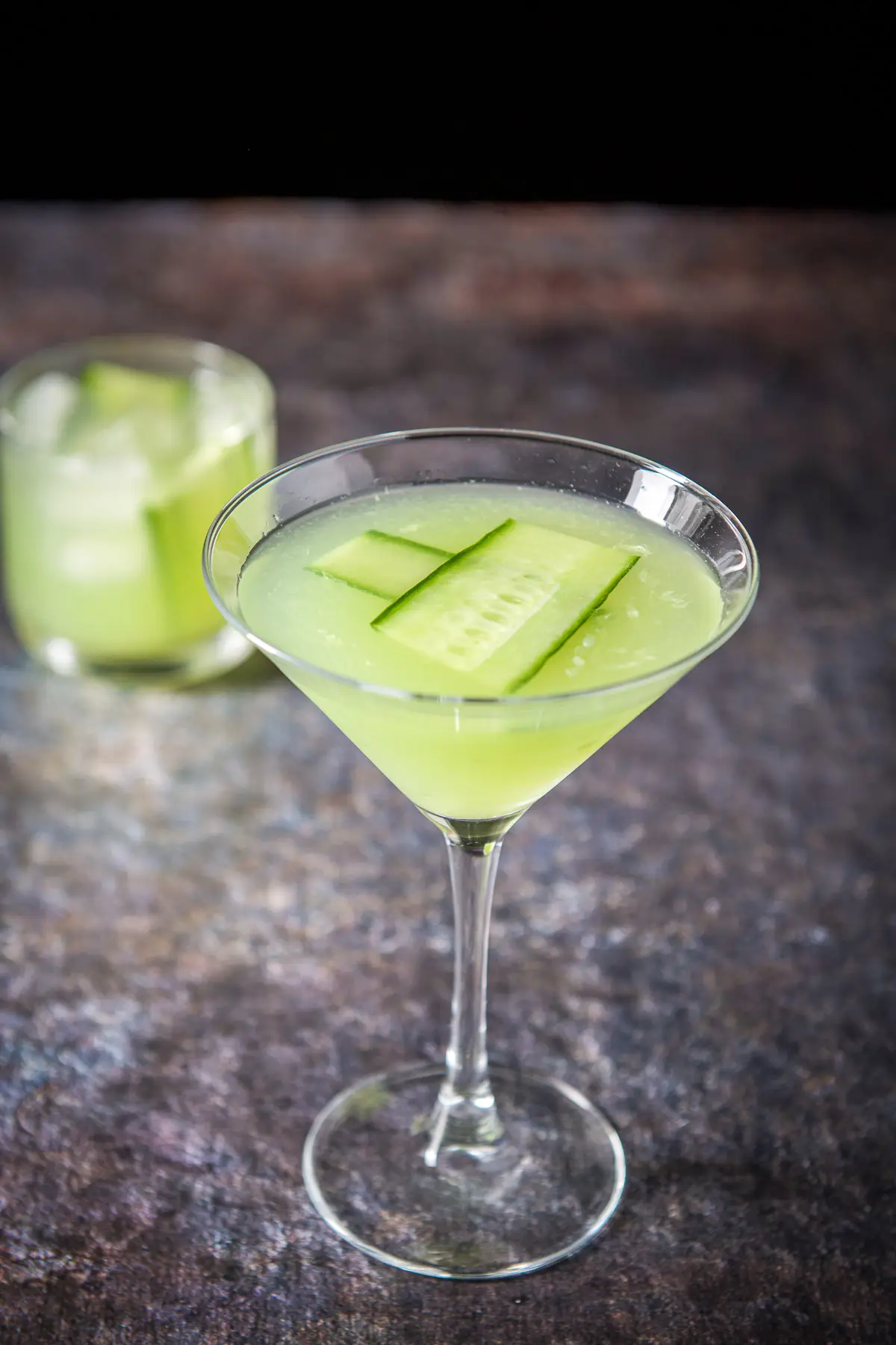 A classic martini glass filled with the cucumber cocktail with a double old fashioned glass in the back