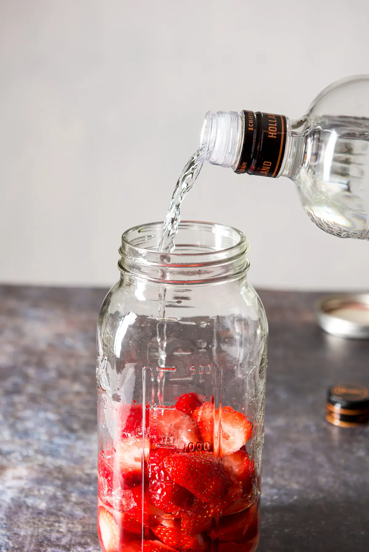 A jar with cut strawberries filled halfway and a bottle of vodka being poured into the jar