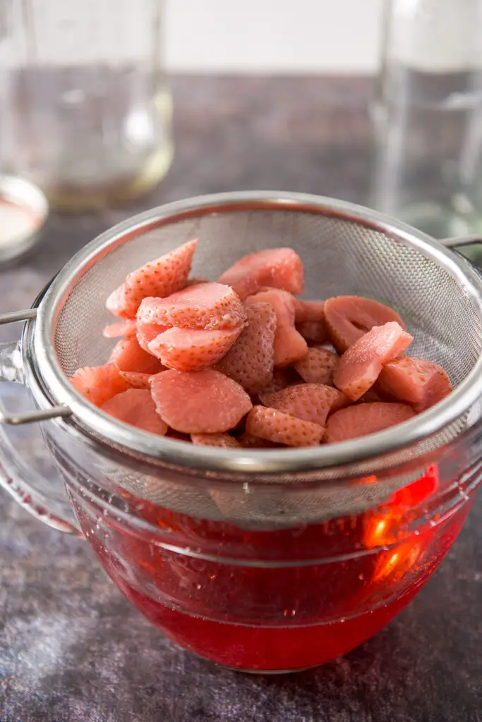 A strainer with pale strawberries in a bowl with the red vodka in it