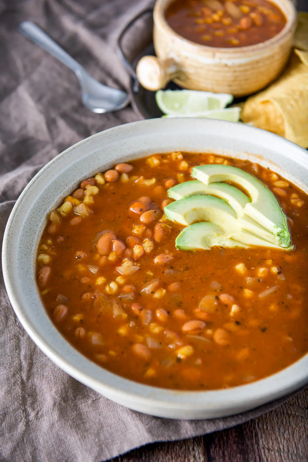 A big bowl with the bean and hominy soup with sliced avocados on top and a crock in the back