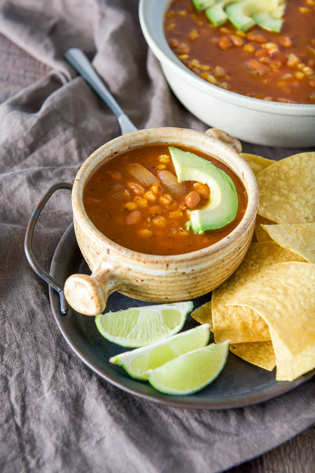 A tray with chips, lime wedges and a crock of soup with an avocado slice on top
