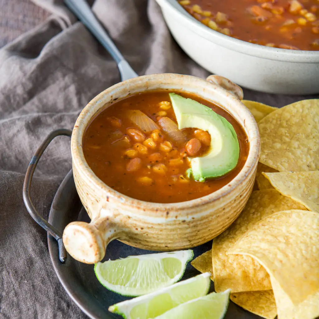 A tray with chips, lime wedges and pozole in a crock with a slice of avocado on it