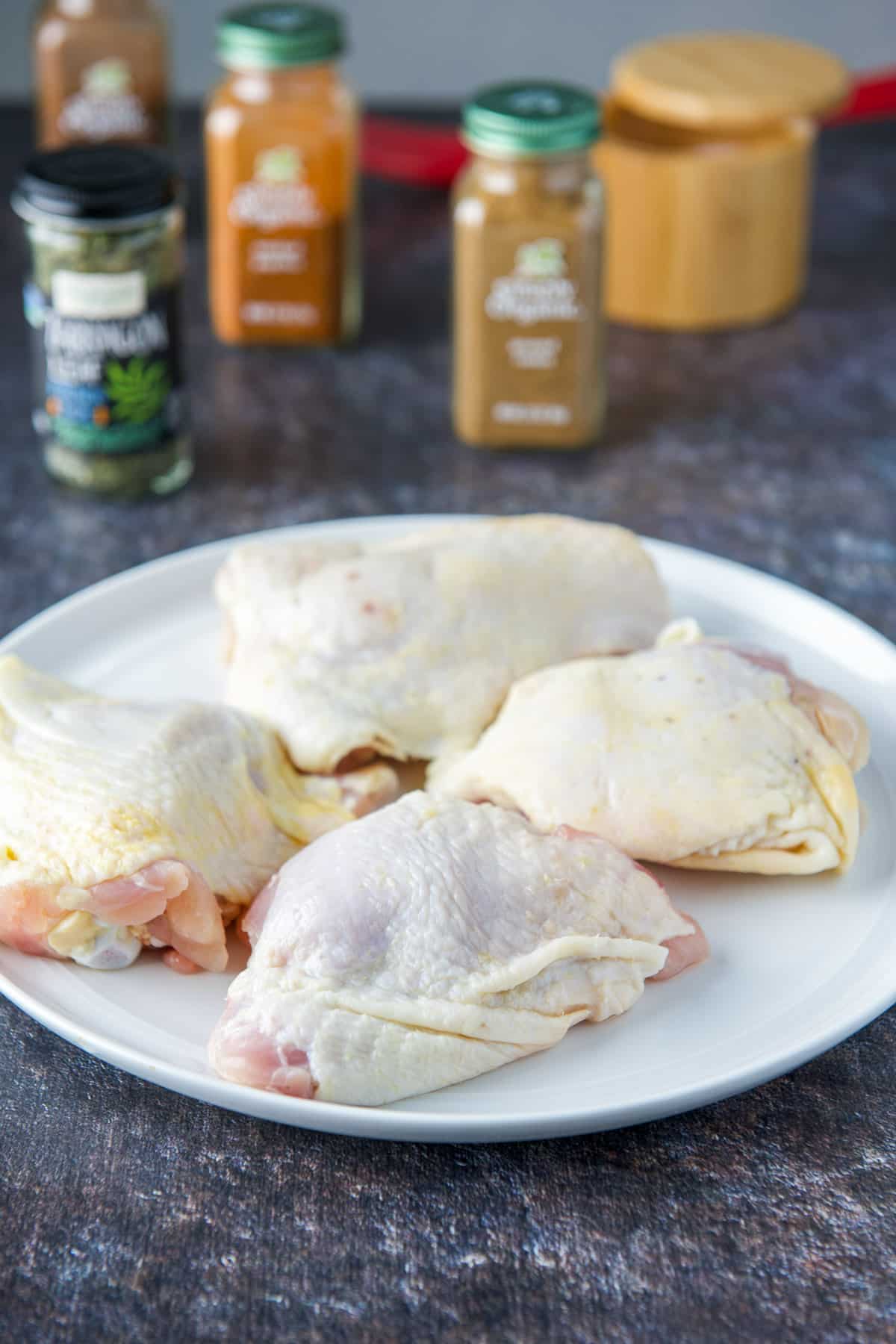 A white plate with raw chicken thighs with spices in the background